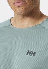 Světle modré sportovní tričko s horizontálními pruhy, kulatým výstřihem a černým logem na hrudi. Materiál vypadá měkce a prodyšně.