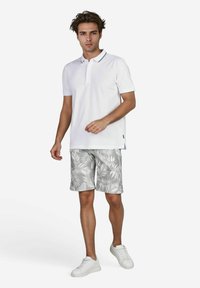 Weißes Poloshirt mit Kragen und einem zweifarbigen Band, kombiniert mit grauen Shorts, die ein weißes Palmenblattmuster aufweisen. Getragen mit weißen Turnschuhen.