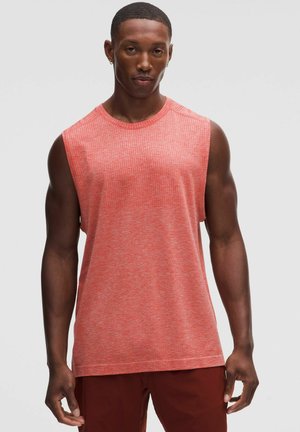 Homme portant un débardeur de sport rouge et un short rouge foncé, debout devant un fond gris clair uni.