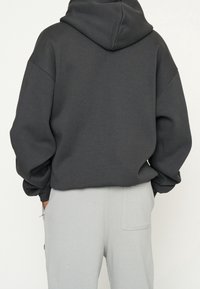 Sweat à capuche gris surdimensionné avec capuche, poignets côtelés et épaules tombantes ; texture douce, associé à un pantalon gris clair avec poches latérales.