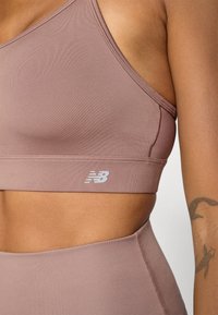 Top desportivo em mauve com um design de costas cruzadas, feito de um material suave e elástico. Apresenta um pequeno logótipo branco "NB" na faixa inferior.