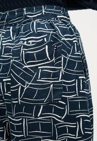 Σκούρο μπλε παντελόνι με άσπρο αφηρημένο μοτίβο γραμμών, ελαστική μέση και πίσω τσέπη με μικρή ετικέτα σε κόκκινο, άσπρο και μαύρο.