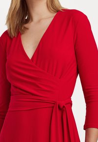 Robe portefeuille rouge avec un profond décolleté en V, des manches trois-quarts et une ceinture nouée à la taille. Tissu lisse avec des détails drapés sur la poitrine.