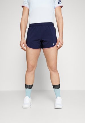 Persoon draagt marineblauwe sportshorts met roze rand, lichtblauwe en zwarte sokken, witte sneakers en een wit shirt met roze rand.