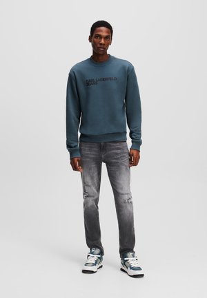Sweat-shirt bleu avec "KARL LAGERFELD JEANS" en lettres noires, associé à un jean gris déchiré et des baskets bleu et blanc.