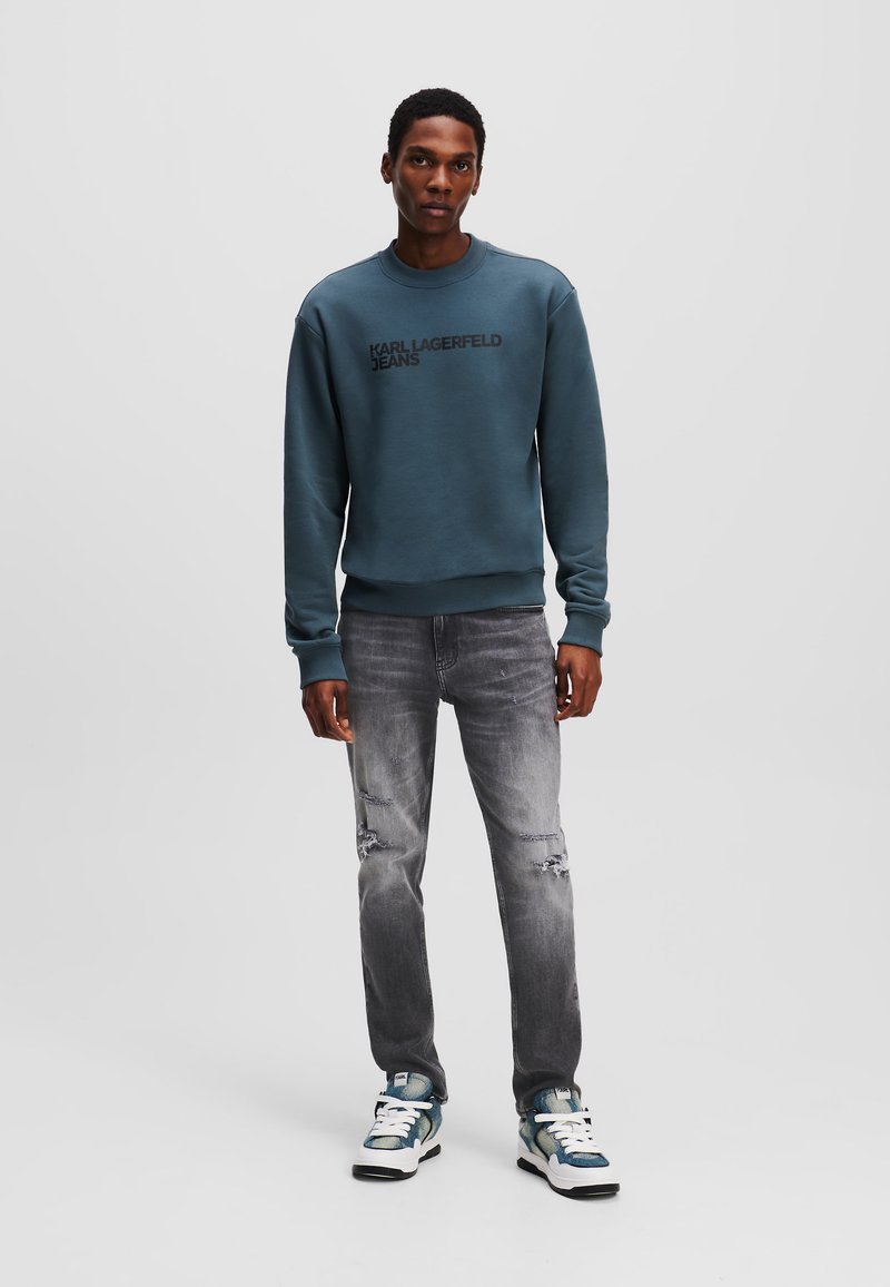 Sudadera azul con "KARL LAGERFELD JEANS" en letras negras, combinada con jeans grises desgastados y zapatillas azules y blancas.