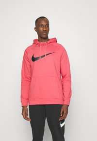 Pink hoodie with a black Nike logo, featuring a front pocket, drawstring hood, and a textured fabric, paired with dark trousers.