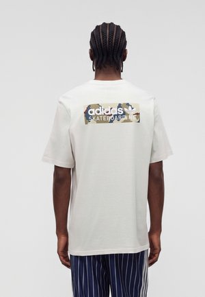 Άτομο με πλεξούδες, που φοράει λευκό T-shirt Adidas Skateboarding και σκούρο μπλε ριγέ παντελόνια, εμφανιζόμενο από πίσω.