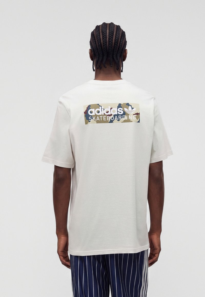 Άτομο με πλεξούδες, που φοράει λευκό T-shirt Adidas Skateboarding και σκούρο μπλε ριγέ παντελόνια, εμφανιζόμενο από πίσω.
