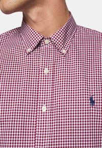 Kostkovaná červeno-bílá košile s límečkem na knoflíky, bílými knoflíky a vyšitou modrou značkou. Bavlněná látka s hladkou strukturou.