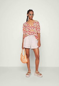 Blusa com padrão floral em rosa e amarelo, calções cinza claro, bolsa de rede laranja e sandálias grossas com tiras leves.