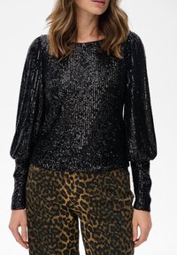 Vrouw in een zwart lange mouwen pailletten top met pofmouwen en hoge taille bruine luipaardafdruk broeken.