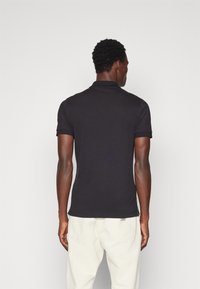 Camisa polo negra de manga corta, hecha de una tela suave, con un cuello doblado y un logo sutil en el dobladillo, llevada con pantalones de color claro.