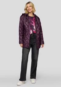 Gesteppte lila Jacke mit Kapuze, ausgestattet mit Reißverschluss und Knopfverschluss. Darüber ein mehrfarbiges Oberteil getragen, kombiniert mit dunklen Jeans und weißen Schuhen.