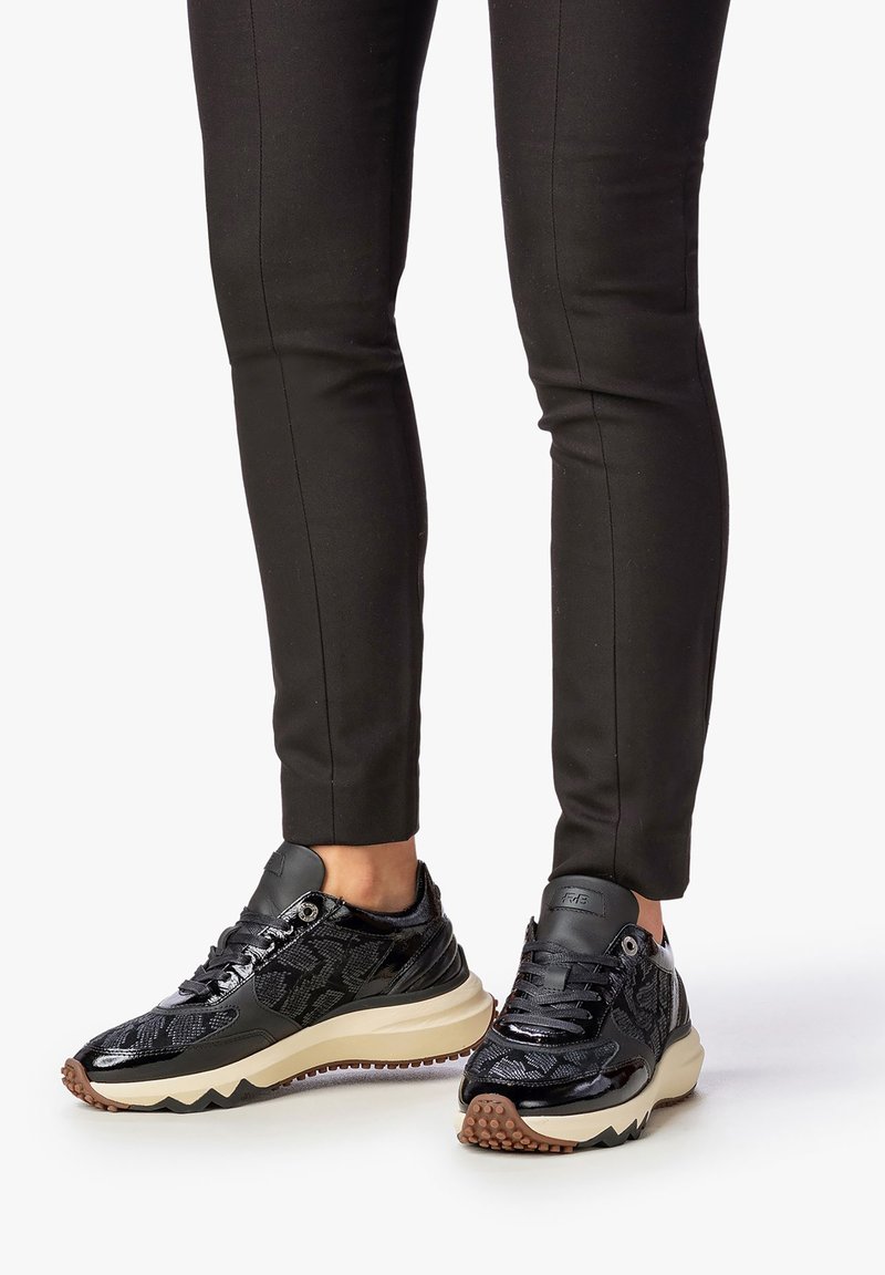 Schwarze Sportschuhe mit strukturiertem Finish, glänzenden Akzenten und einer klobigen, cremefarbenen Sohle, kombiniert mit enganliegenden schwarzen Hosen.