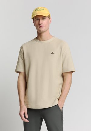 T-shirt beige à manches courtes avec une finition texturée et un col rond. Présente un petit logo hexagonal noir sur la poitrine.
