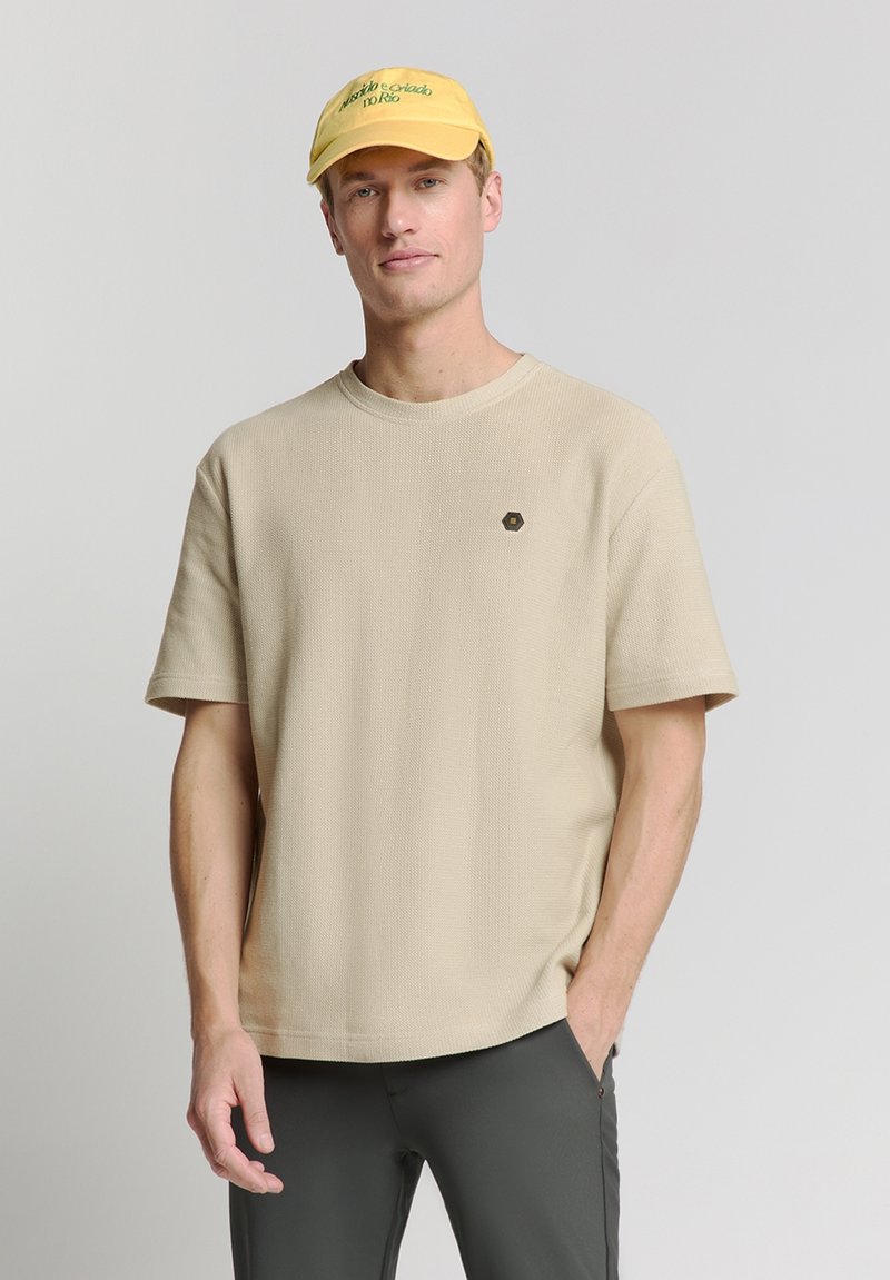 Beige T-shirt met korte mouwen, met een gestructureerde afwerking en een ronde halslijn. Bevat een klein zwart hexagonaal logo op de borst.