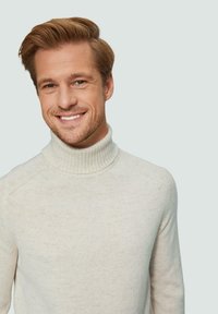 Crème-kleurige turtleneck trui in een ribbelbrei textuur met ingeknipte schouders. Model laat een lichte glimlach zien tegen een lichte achtergrond.