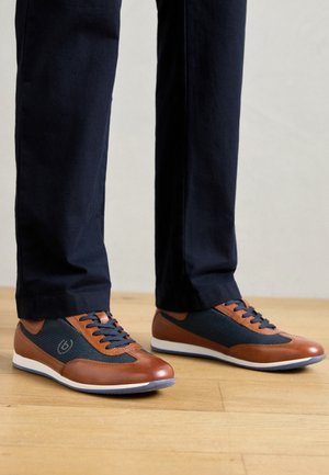 Brune lærred og marineblå mesh-sneakers med blå snører, med en hvit såle og logo detaljer på siden, iført mørke bukser på et tregulv.