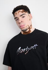 Zwarte katoenen T-shirt met een glanzend, metallic logo op de borst. Korte mouwen en een ronde halslijn, met een relaxed fit.