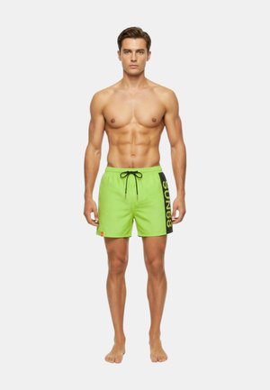 Giovane uomo senza maglietta, in piedi a piedi nudi, che indossa pantaloncini da bagno verde acceso con la scritta "SUN68" stampata sul lato, su uno sfondo bianco semplice.