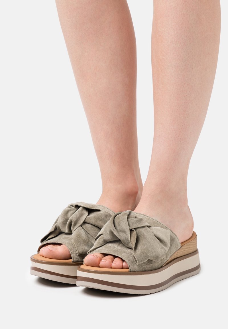 Pieds portant des sandales slides beiges avec des semelles compensées épaisses et de larges brides en tissu nouées sur les orteils.