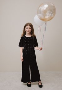 Tuta in velluto nero con motivo a cuori, maniche corte e design a gamba larga. Il bambino tiene due palloncini con nastri pastello.