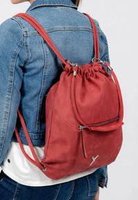 Sac à dos à cordon en cuir rouge perforé avec bretelles ajustables, poche avant zippée et accessoires en argent. Finition texturée.