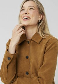 Manteau boutonné marron avec un col, en tissu doux et texturé, et boutons noirs. Le modèle touche doucement son menton en souriant.
