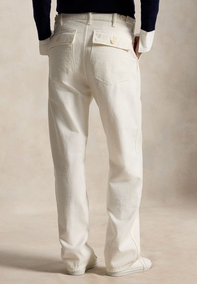 Persoană purtând pantaloni drepți de culoare off-white cu buzunare aplicate cu clapă în spate și detalii cu nasturi, mâinile în buzunarele laterale, fundal neutru.