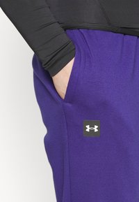 Lila sweatpants med en slät textur, som har ett svart logopatch, sidofickor och en elastisk midja. Lång svart åtsittande ärm synlig.