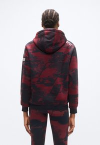 Sweat à capuche avec un motif géométrique rouge foncé et noir. Confectionné dans un tissu doux, il présente des poignets côtelés et un logo sur l'épaule.