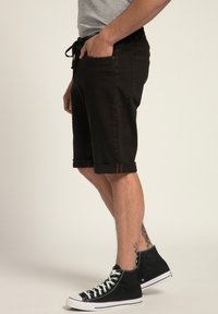 Shorts in denim nero con vestibilità comoda, caratterizzati da orli arrotolati e cintura con cordino. Indossati con scarpe da ginnastica alte nere.