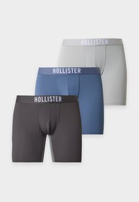 Trois paires de boxers Hollister en gris, bleu et noir, empilées en arrangement décalé sur fond blanc.