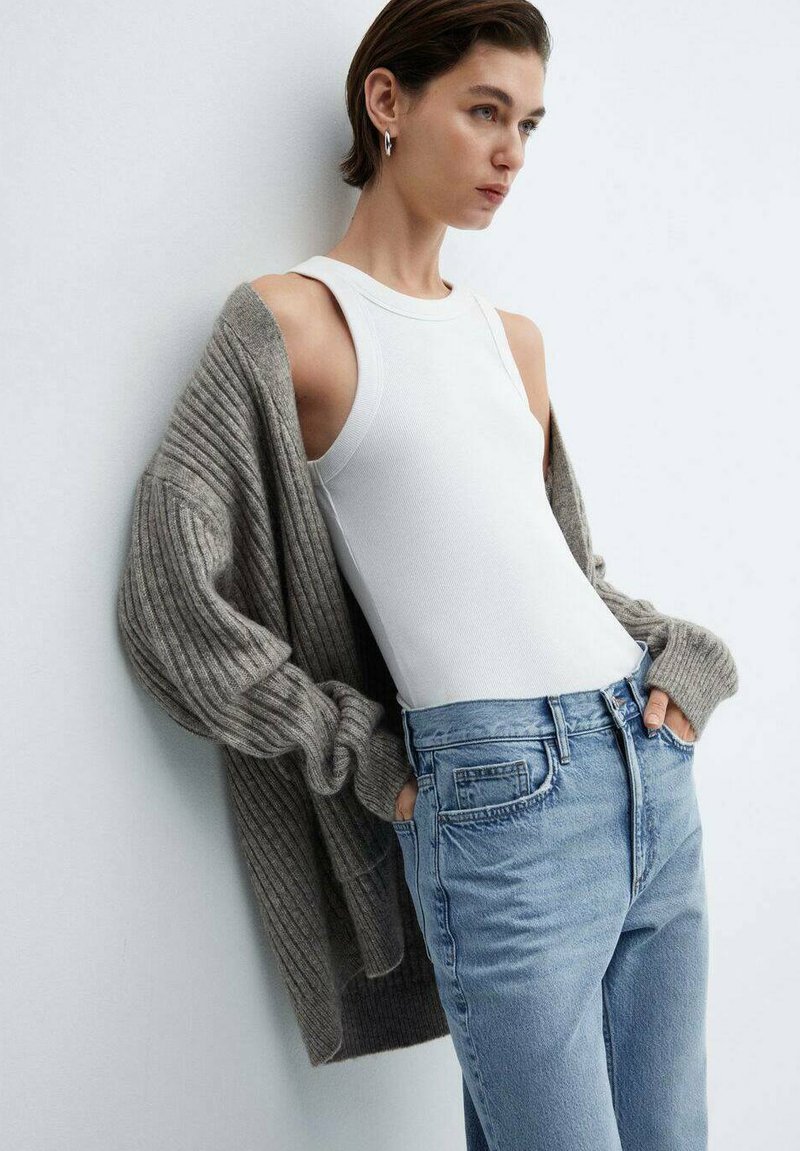Weißes Tanktop kombiniert mit einem weichen, gerippten grauen Cardigan. Das Model trägt hellblaue High-Waist-Jeans mit Taschen. Schlichter, lässiger Look.