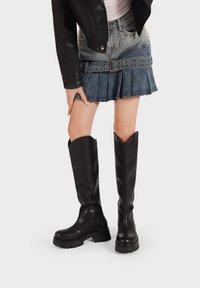 Stivali neri fino al ginocchio in pelle liscia, con una suola spessa, una parte superiore angolare e un design minimalista. Indossati con una gonna di jeans.