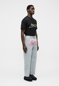 Μαύρο μπλουζάκι με γραφικό σχέδιο, ανοιχτό μπλε τζιν παντελόνι με ροζ εκτύπωση καρτούν γάτας και σκούρα παπούτσια slip-on. Καθημερινή εφαρμογή, χωρίς ορατά λογότυπα.