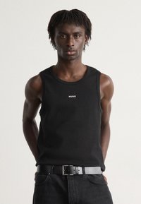 Un maiou negru fără mâneci, cu un guler rotund și un mic logo alb "HUGO". Asortat cu o curea neagră și blugi închiși la culoare.