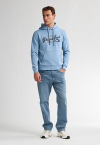 Sudadera con capucha de color azul claro con un bolsillo frontal y un logo impreso en negro, combinada con jeans azul claro y zapatillas blancas.