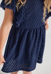 Granatowa sukienka wykonana z teksturowanej tkaniny z wzorami w formie oczek, z krótkimi, falbaniastymi rękawami i zgromadzonym w pasie materiałem.