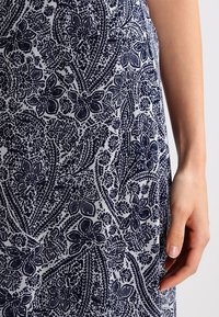 Laivastonsiniseen ja valkoiseen kukkakuviollinen kangas, jossa on yksityiskohtaisia kukkia ja lehtiä teksturoidulla taustalla. Osa kädestä näkyvissä.