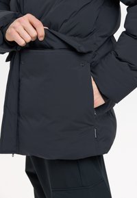 Schwarze gepolsterte Jacke mit einem Drahtkragen, Seitentaschen und vorderen Knöpfen. Der Stoff ist glatt mit einer strukturierten Oberfläche.
