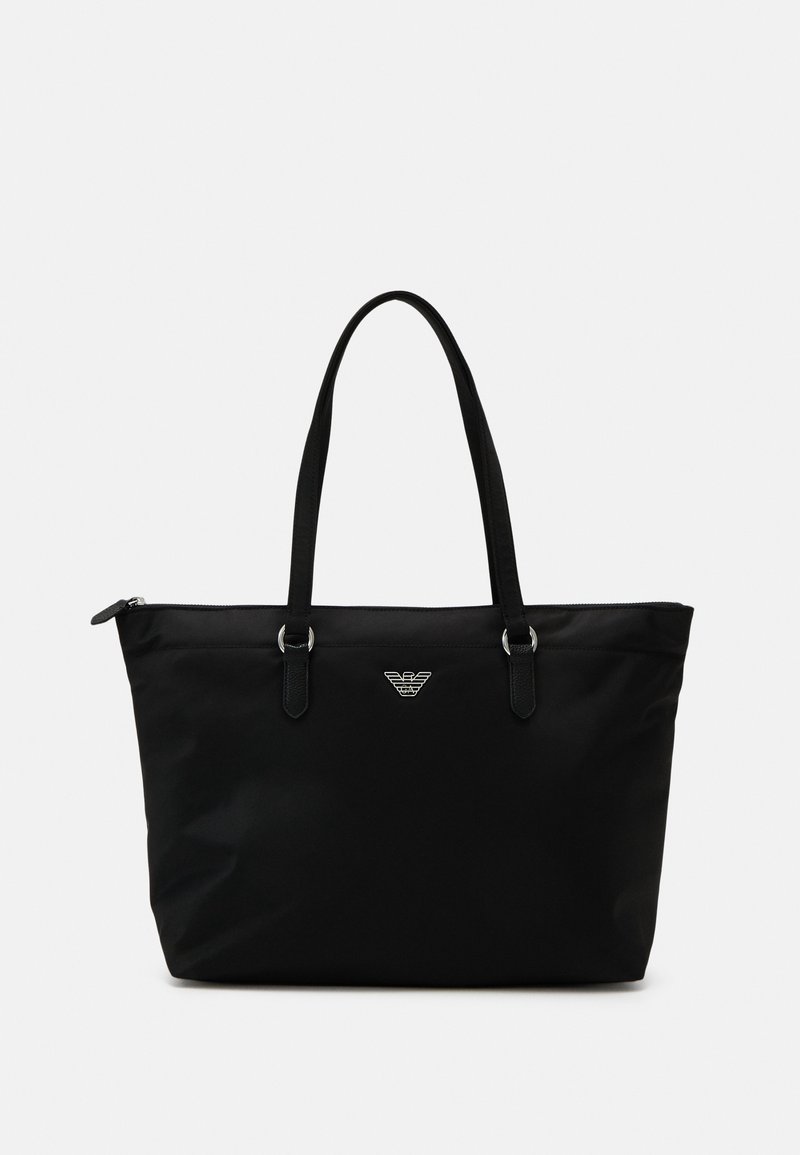 Sac fourre-tout noir avec des poignées longues, emblème logo argenté centré sous la fermeture éclair supérieure, sur un fond clair uni.