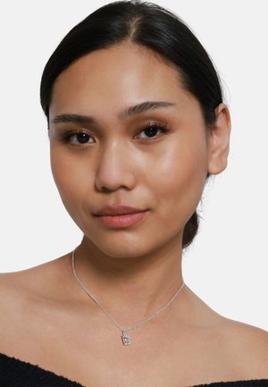 Silberne Kettenhalskette mit einem rechteckigen Anhänger mit strukturierter Oberfläche. Das Modell hat glattes, dunkles Haar und einen neutralen Ausdruck.