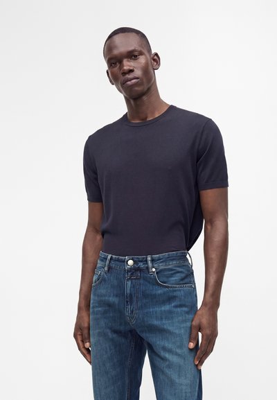 Homme portant un t-shirt noir uni à manches courtes rentré dans un jean en denim bleu, debout sur un fond blanc.