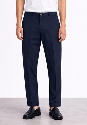 Mężczyzna ubrany w granatowe, dopasowane spodnie ze zwiniętymi nogawkami i czarne skórzane mokasyny, stojący na szarej podłodze na tle białej ściany.