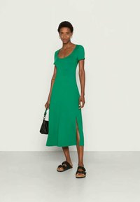 Mulher vestindo um vestido midi verde com fenda lateral, sandálias pretas e segurando uma pequena bolsa preta, em pé contra um fundo branco liso.