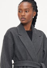 Cappotto grigio in misto lana strutturato con vita arricciata, ampi risvolti e due tasche frontali, indossato sopra una maglia nera.