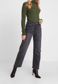 Jean taille haute gris foncé à coupe droite, associé à un pull à col roulé côtelé vert et des bottines noires à bout pointu.