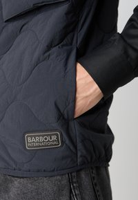 Jachetă cu quilting în culoare bleumarin, cu un patch din cauciuc, buzunar lateral și manșete negre, realizată dintr-un material ușor.